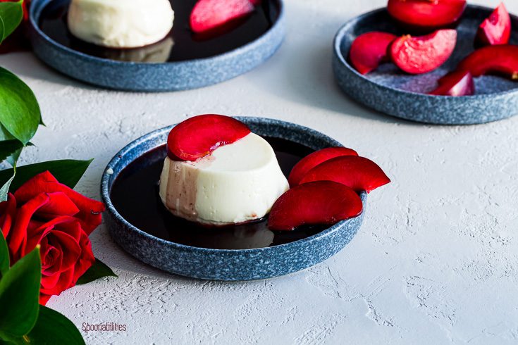 Three blue plates: one in the front with panna cotta and red wine sauce with fruits, the plate in the right side with slices of plumcots and the third one in the left side with panna conna. Spoonabilities.com