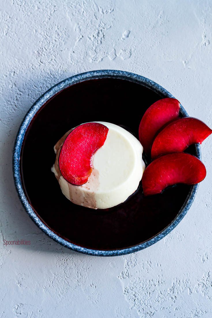 Labneh panna cotta in a round blue plate from anfora. The panna cotta is garnished with slices of plumcots and a red wine syrup. Spoonabilities.com