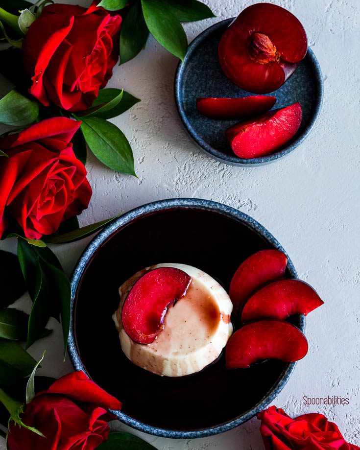 Panna Cotta with red wine syrup in a round blue plate with red roses around the plate with another plate in the uppper side of the photo with plumcots slices. Spoonabilities.com