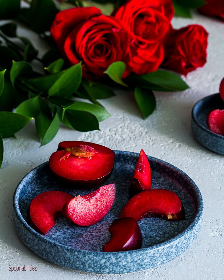 Red Plumcots slices in a round blue plate with red roses in the background. Spoonabilities.com