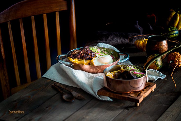 Wooden tabletop decorated with Fall vibes and mini pumpkins. In the center of the table a personal two handle fry pan with Brown Butter Mashed Pumpkin-Cauliflower, braised short ribs, fry egg, microgreens and pickled onions, Spoonabilities.com