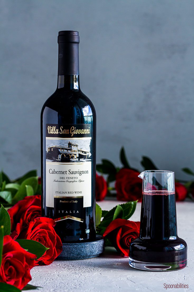 A bottle of Cabernet Sauvignon from Villas San Giovanni on top of a small blue saucer with red roses around the wine. In another small carafe the red wine syrup. Spoonabilities.com