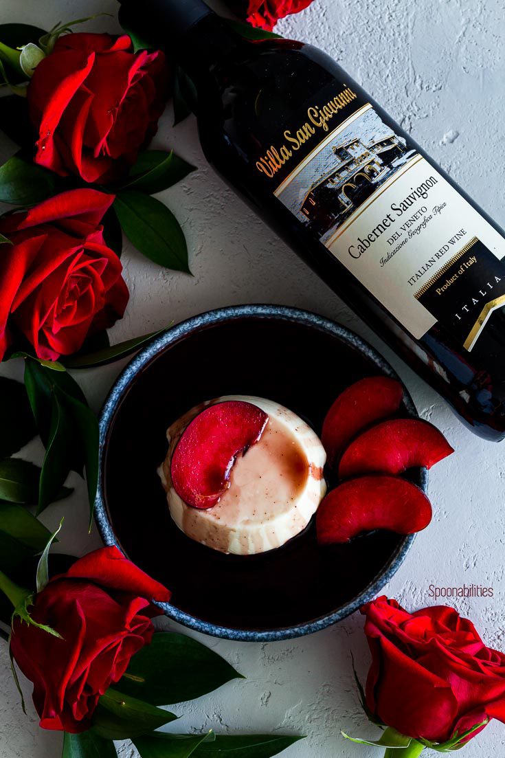 Lay flat photo with panna cotta in a round blue plate surrounding red roses and a bottle of Villas San Giovanni Cabernet Sauvignon. Spoonabilities.com