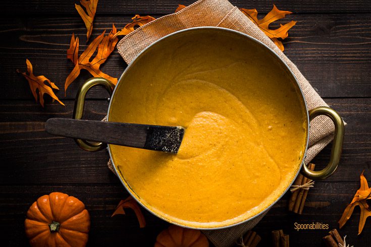 Pot with the Dominican arepa mixture after have been cooked in the stove with a spoon inside the pot. The background is decorated with Mini pumpkin, leaves & cinnamon sticks. Spoonabilities.com