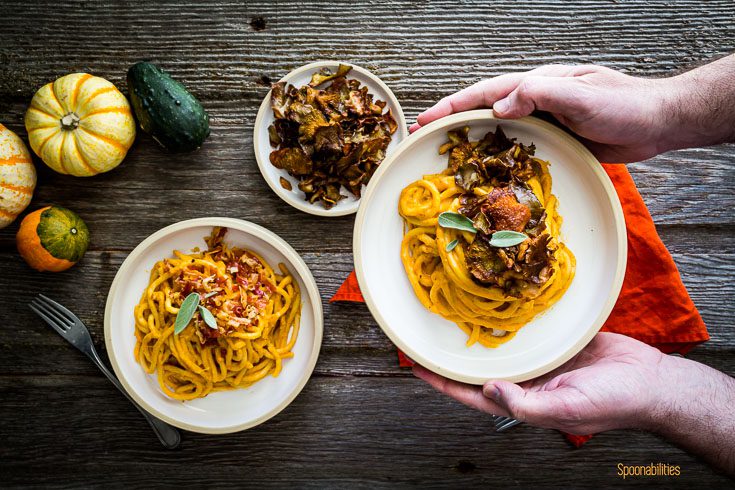 Two hands holding a bowl with pasta coated in creamy pumpkin pasta sauce and topped with crispy oyster mushrooms. two more plates on the far left of the table. Spoonabilities.com