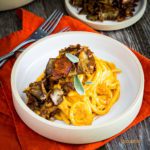 Tuscany spaghetti pici pasta coated in pumpkin pasta sauce with crispy mushrooms in a round plate on top of a orange napkins