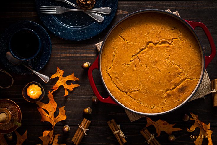 Dominican Arepa in a red cast iron casserole after came out of the oven. The scene is decorated with bluer plates, cups and flatware with orange leaves around the table with a small candle on the left side. Spoonabilities.com