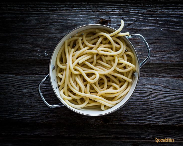 Cooked pici pasta in a white colander on a wooden surface. Spoonabilities.com