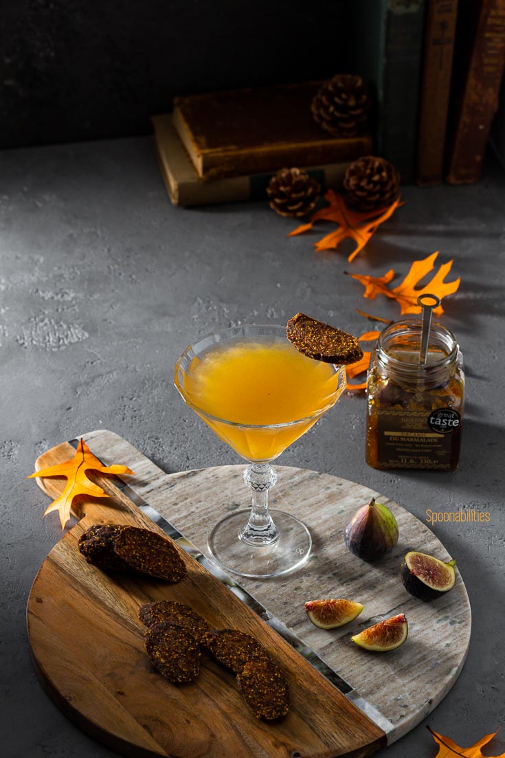 Fig Martini in a beautiful glass on a serving board. Next to the board a jar of fig marmalade. The acacia marble board is garnished with fresh fig and slices of vegan fig salami. Spoonabilities.com