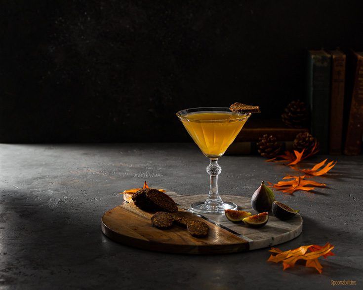 Horizontal photo with a beautiful Vintage cocktail glass on a round board with a Fig martini made with fig jam and vodka. Garnished with a slice Vegan Fig Salami. The marble acacia board has fresh fig and a slices of the vegan fig salami.