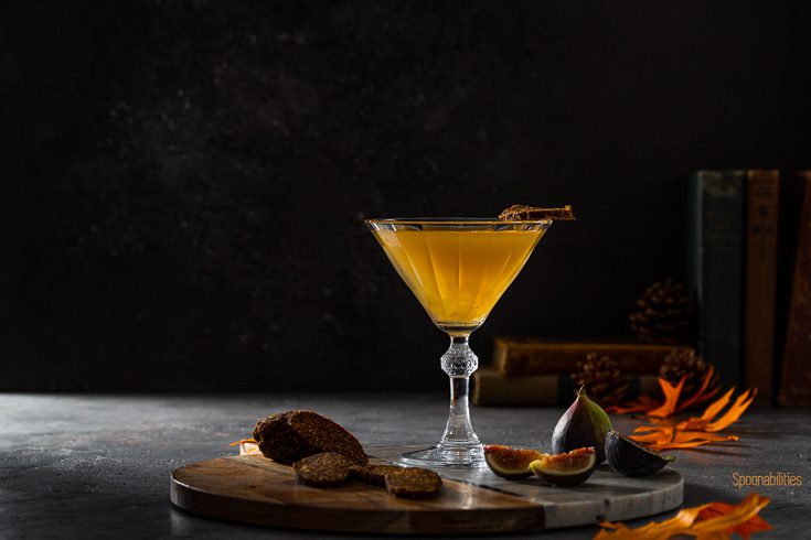 Vintage cocktail glass on a wooden/marble board with a delicious martini made with fig preserves/jam from Navarino Icons and vodka. Garnished with a slice Vegan Fig Salami with Orange. The board has cut fresh fig and a slices of the vegan fig salami. Available at Spoonabilities.com