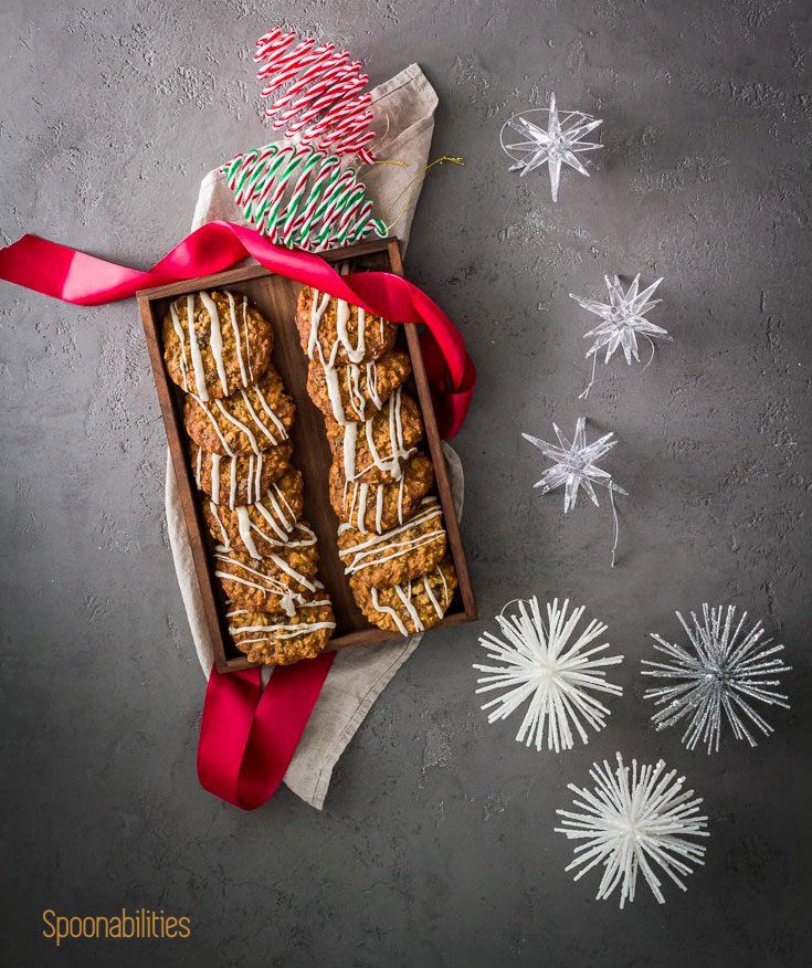 Flat lay photo with a wooden tray with Oatmeal Raisin Maple Bourbon Cookies with a red ribbon and holiday decoration on the left side of the photo. Spoonabilities.com