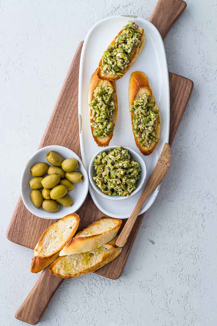 Overhead shot of 3 Greek Olive Crostini on a white plate next to green olives and baguette slices