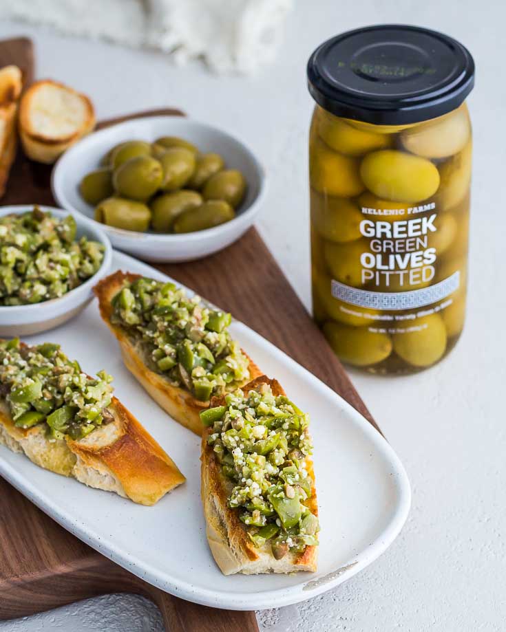 Three servings of Greek Olive Crostini on a white plate next to a jar of Greek Green Olives