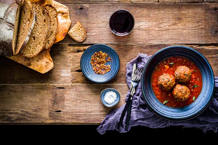 Overhead photo with one bowl with three Jumbo Brisket Meatballs in San Marzano tomato sauce. Next to the bowl two small saucer one with sea salt and the second one with smoked chili pepper and in the right top corner a loaf of bread. Spoonabilities.com