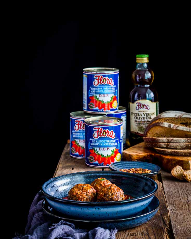 Blue bowl in the center with meatballs and in the background 4 cans of San Marzano D.O.P. Tomatoes and a glass bottle with olive oil from Flora Fine Foods. Spoonabilities.com