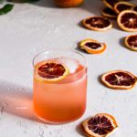 Blood orange vodka soda with a trail of dried blood orange slices on a gray background