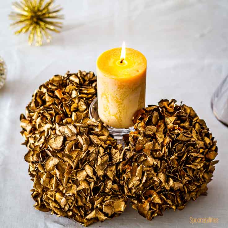 A pillar candle in a base with golden flowers on a table in a white tablecloth. Spoonabilities.com