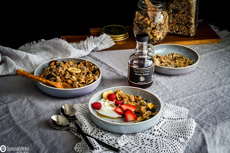 Table presentation with three bowls. One in the front with yogurt, easy no-grain granola with strawberry, raspberry and a drizzle of maple syrup. The back bowls have granola and in betwen the bowls there is a jar of maple syrup which is available at Spoonabilities.com