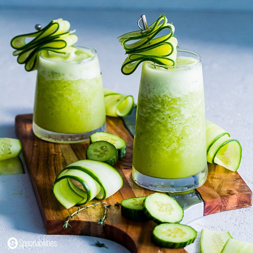 Two glasses on a wooden board with cucumber margarita slush. Garnished with shaved cucumbers. Recipe at Spoonabilities.com