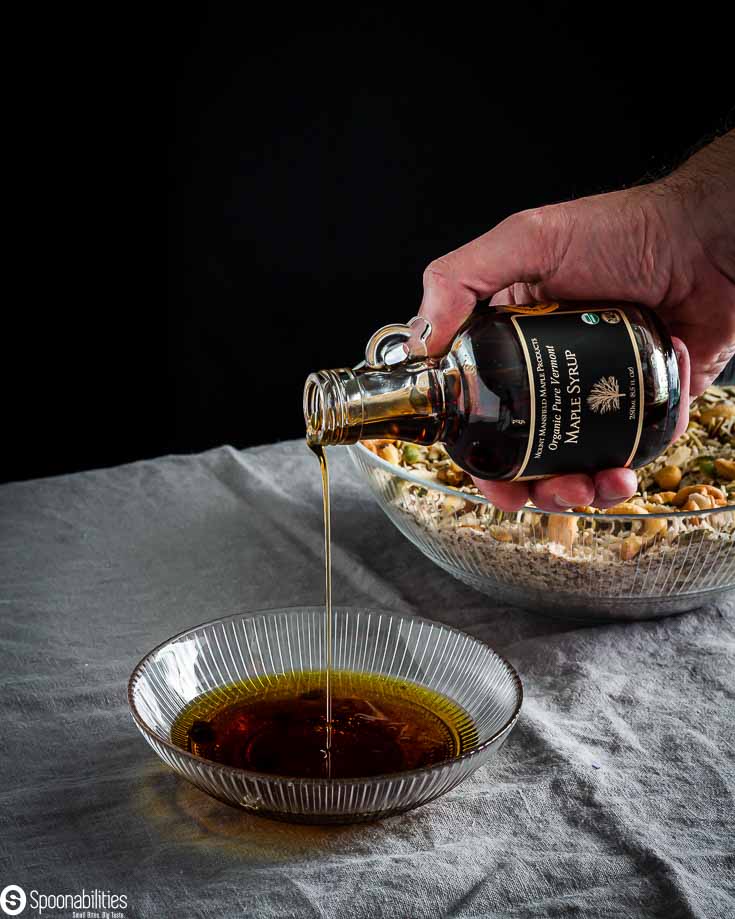 Small glass bowl the wet ingredients for the easy granola recipe with Greek extra virgin olive oil and a hand pouring maple syrup. Both products available at Spoonabilities.com