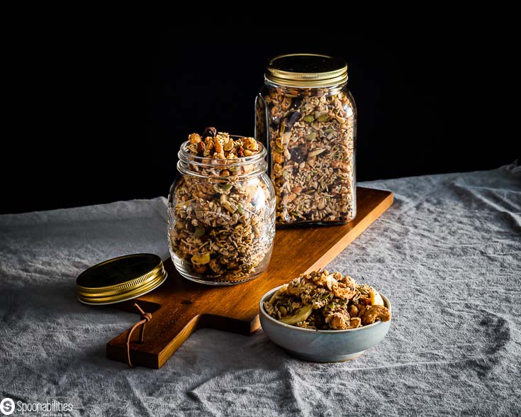 Granola packed in two glass jars on top of a wooden board and next to the front jar, there is a small bowl with some granola. Available at Spoonabilities.com