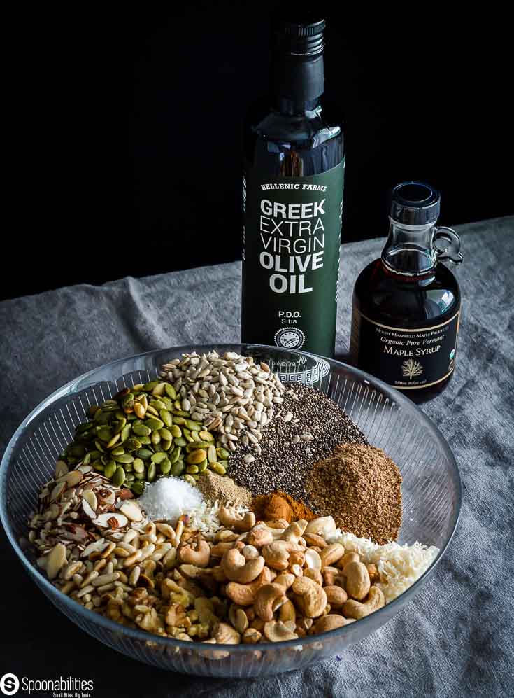 A large glass mixing bowl with pantry staples food like nuts, seeds, spices, coconut flakes. And, next to the bowl a bottle of Greek EVOO and maple syrup, both products available at Spoonabilities.com