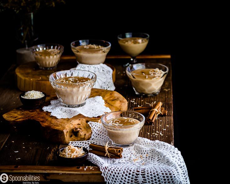 Table presentation with 6 dessert glass bowl with Jalea de Batata Dulce on top of a wooden table. The front glass bowl is on top of a small napkin with a couple of cinnamon stick and toasted coconut flakes near by. See recipe at Spoonabilities.com