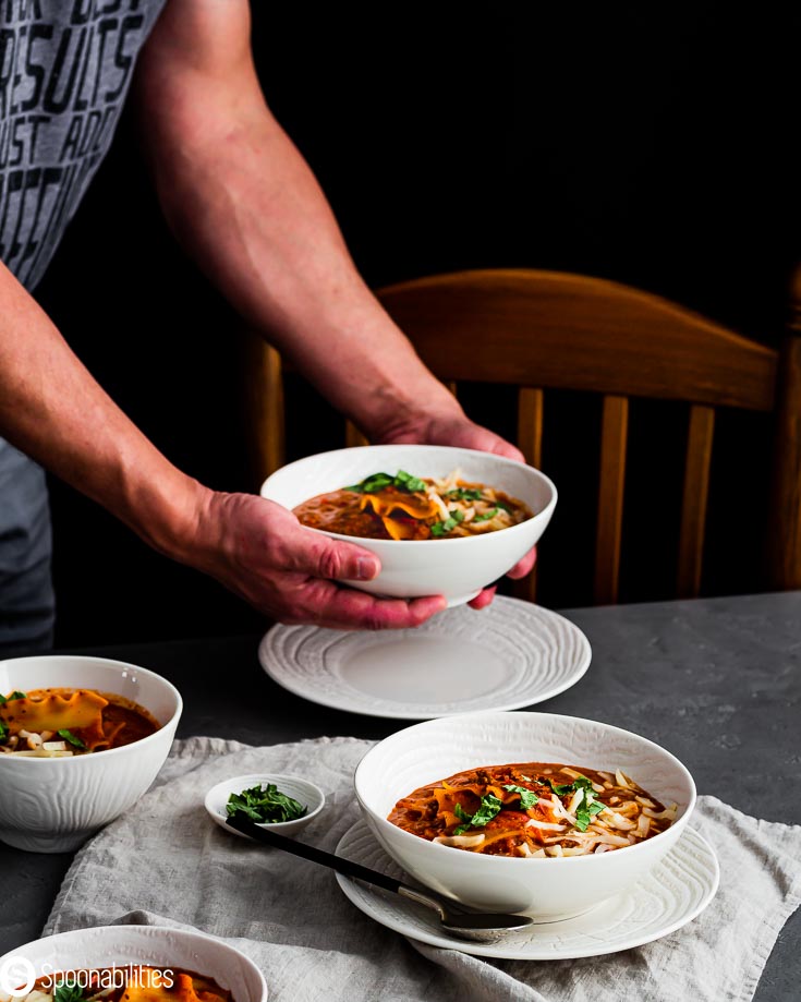 Table with three white bowl with lasagna soup and a fourth one in my hands putting the bowl on the table. Spoonabilities.com