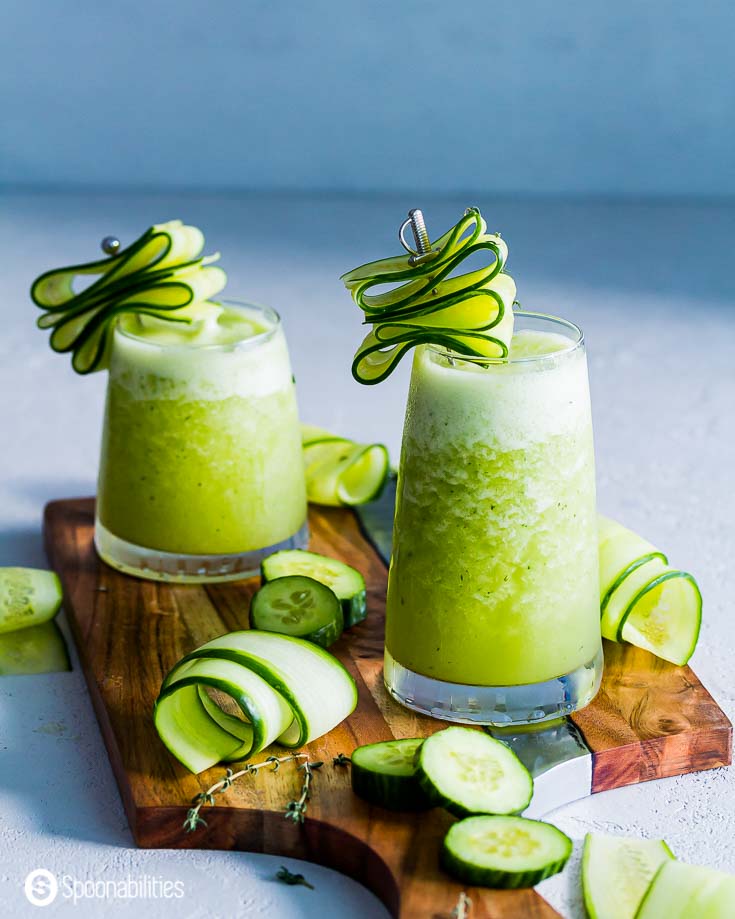 Two glasses on a wooden board with cucumber margarita slush. Garnished with shaved & sliced cucumbers. Recipe at Spoonabilities.com
