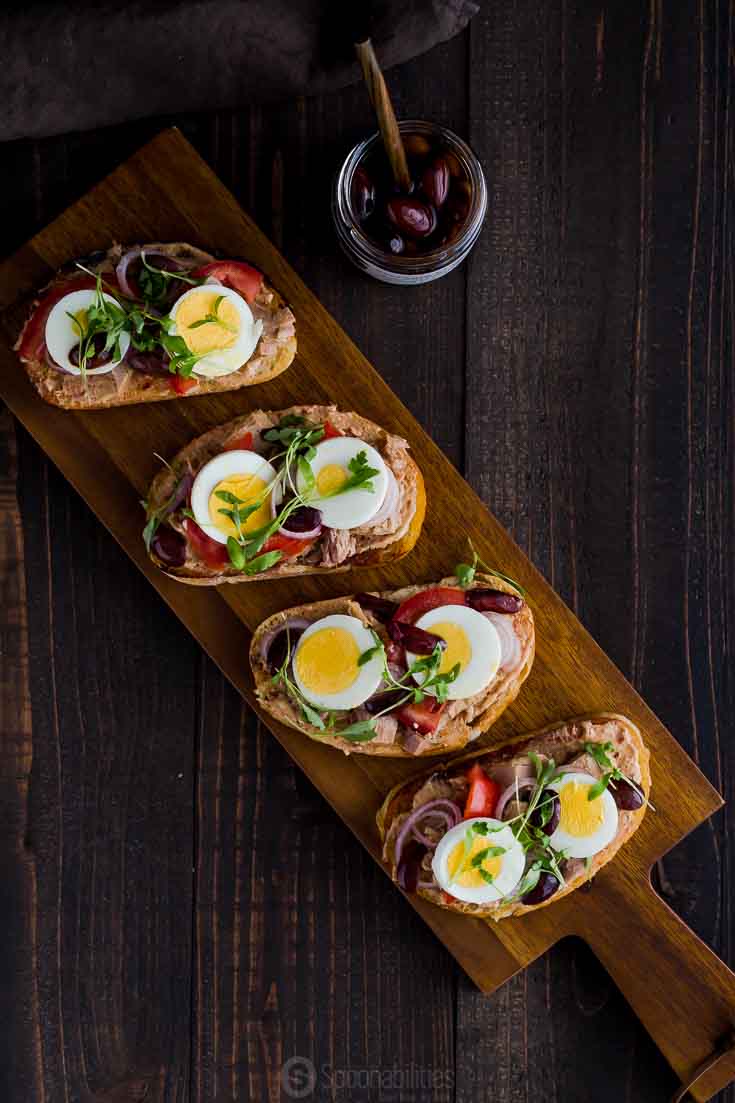 Overhead photo with a wooden serving board with four tuna toast with eggs, tomato and kalamata olives. Available at Spoonabilities.com