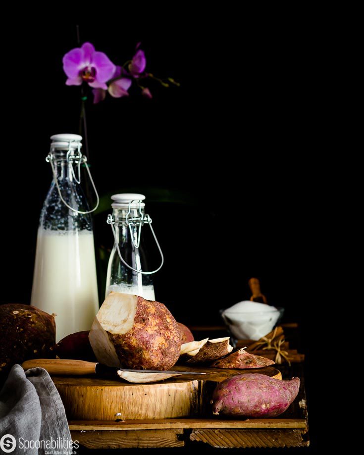 Ingredients for the White Sweet potato pudding: white sweet potato on a cutting board getting peal, on the left side two milk bottle one with coconut milk and the other one with evaporated milk and in the background a glass bowl with sugar. Recipe at Spoonabilities.com