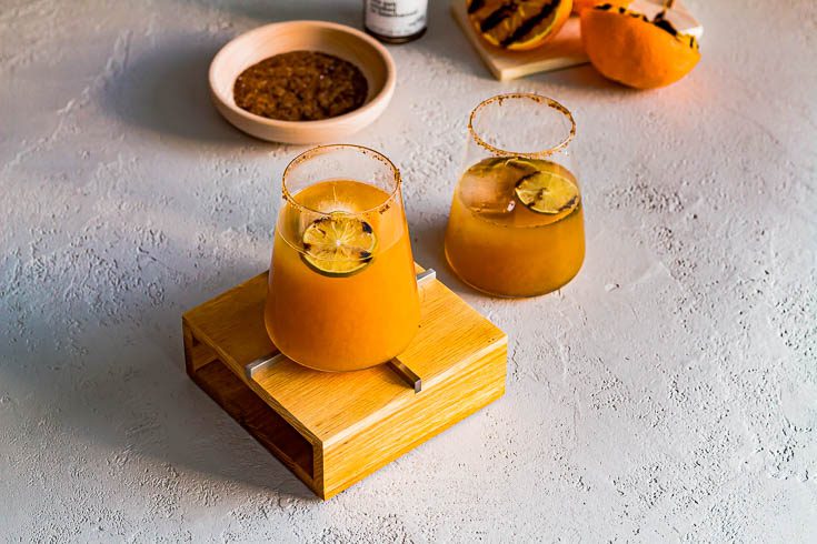 Two tumbler with Grilled Orange Margarita, one of the tumblers is on top of a wooden riser. In the back of the photo is wooden plate with the smoked sea salt. Recipe at Spoonabilities.com