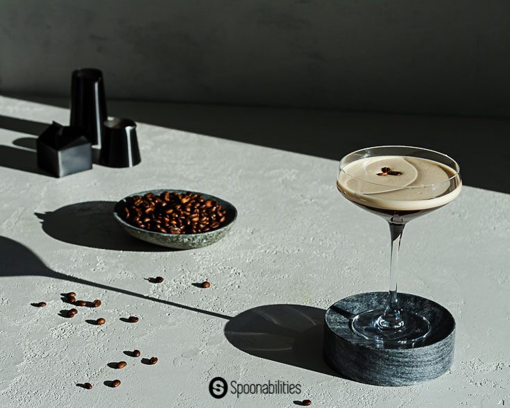 Espresso martini on a dark gray pedestal with long shadow and plate of coffee beans