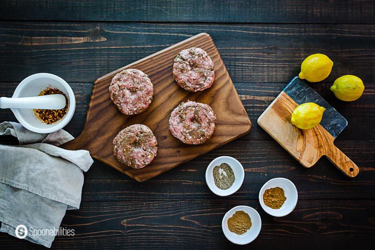 Four spiced lamb patties in a wooden peel board. In a smaller peel board 3 lemons and some pinch bowls with spices around the table. Recipe at Spoonabilities.com