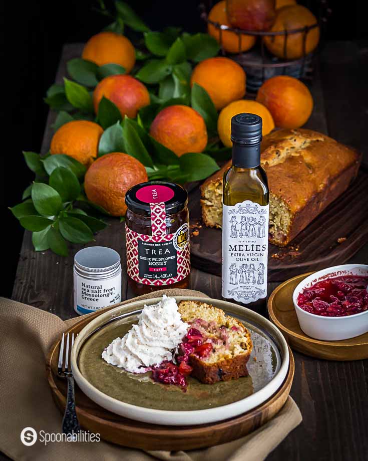 Blood Orange olive oil cake in the front of the photo on a plate. In the background Melies extra olive oil, Trea Wildflower honey & Greek Sea Salt. More gourmet products at Spoonabilities.com