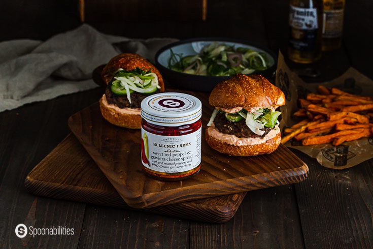 A jar of Sweet Red Pepper & Graviera Cheese Spread. In the background two spiced lamb burger with sweet potatoes fries on the side. Spoonabilities.com