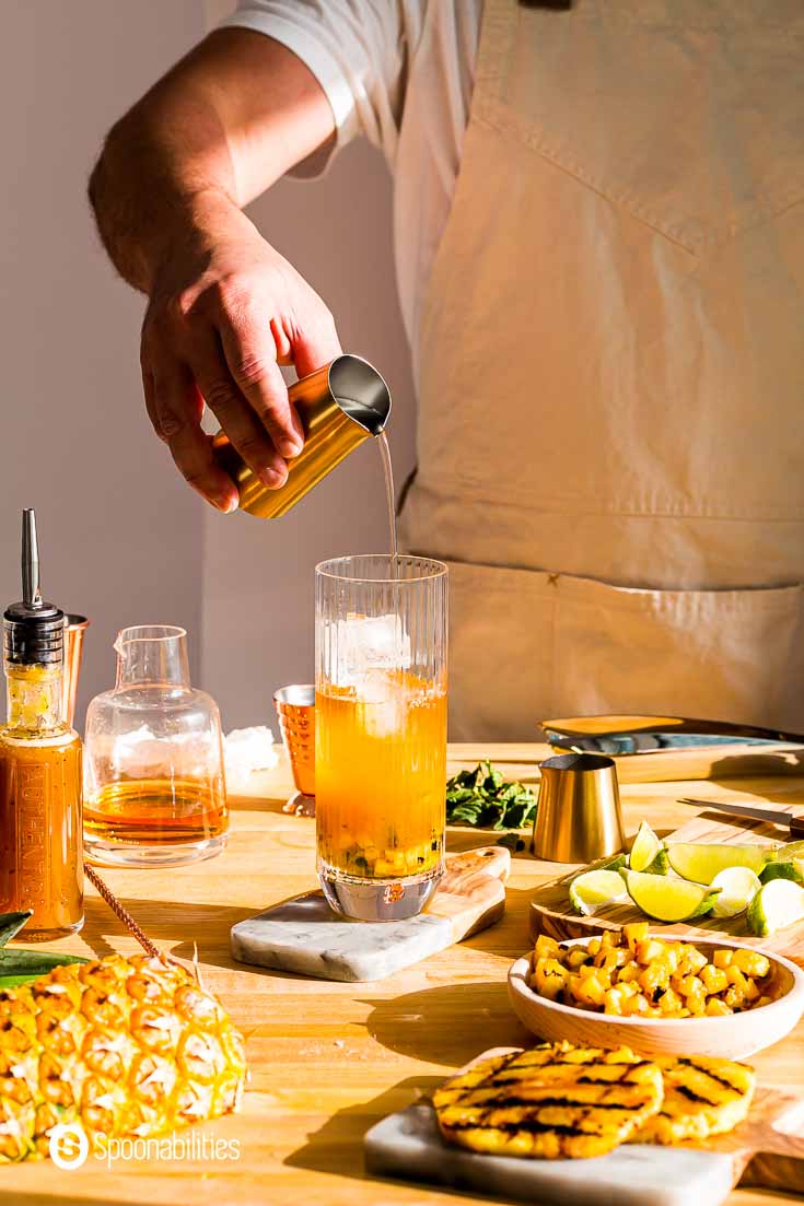 Preparing the grilled pineapple mojito. The glass is in the center on top of a small cutting board. And, around the glass bar tools and pineapple pieces and lime wedges. Recipe at Spoonabilities.com