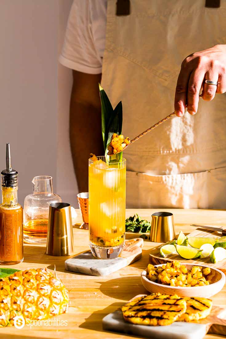 Garnishing the grilled pineapple mojito with pineapple chunks. The glass is in the center of the photo on top of a small wooden cutting board. Recipe at Spoonabilities.com