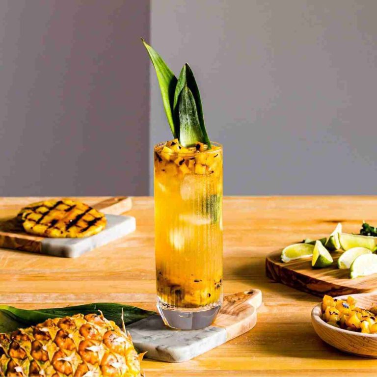 A tall glass in the center of the photo with a Grilled Pineapple Mojito. Around the glass pieces of pineapple and cutting board. Recipe at Spoonabilities.com