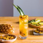A tall glass in the center of the photo with a Grilled Pineapple Mojito. Around the glass pieces of pineapple and cutting board. Recipe at Spoonabilities.com