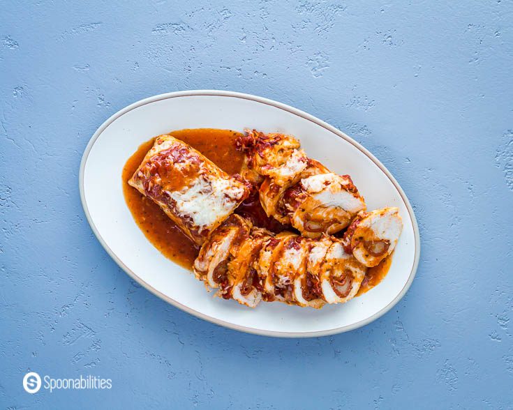 Overhead photo of the stuffed pizza chicken, served in a oval plate. Some of the pieces of the chicken are showing the filling in the center and there is another piece without cutting. Recipe at Spoonabilities.com