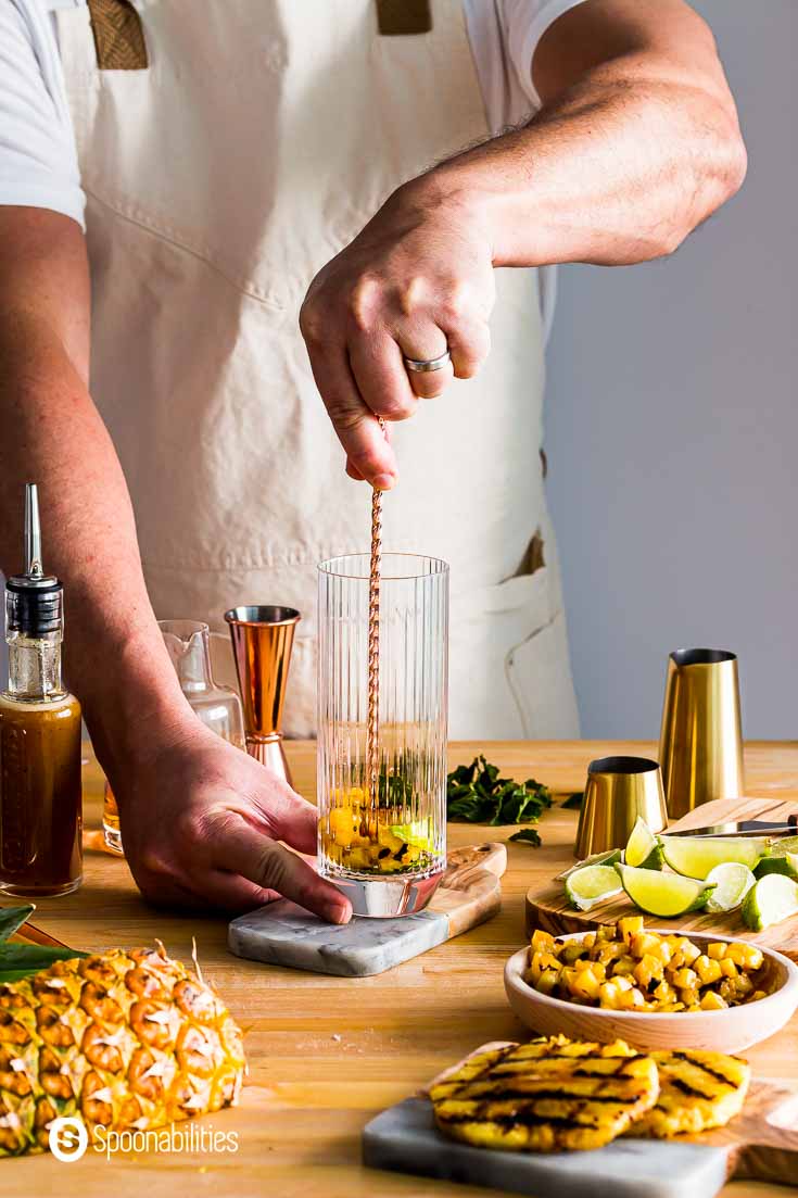 A guy muddling and smashing away lime juice, grilled pineapple chunks, and mint. The glass is in the center of the photo and around the glass chunk & rings of grilled pineapple, lime wedges and bar tools. Recipe at Spoonabilities.com