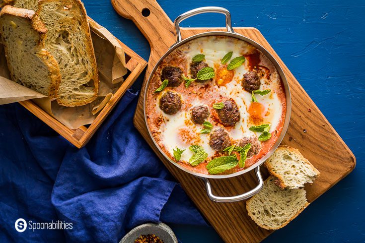 Blue background with a small saucepan with eight lamb meatballs in a tomato & red pepper sauce with three poached eggs. Served with 3 crusty bread in a wooden basket and smoked chili flakes in a small stone bowl. Recipe at Spoonabilities.com