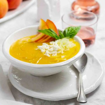 A table with white tablecloth, white plates and bowls, and on the right side a tumbler with rose wine. In the bowl there is a Chilled Peach Soup with Fresh Goat Cheese. Recipe at Spoonabilities.com