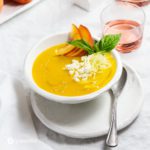 A table with white tablecloth, white plates and bowls, and on the right side a tumbler with rose wine. In the bowl there is a Chilled Peach Soup with Fresh Goat Cheese. Recipe at Spoonabilities.com