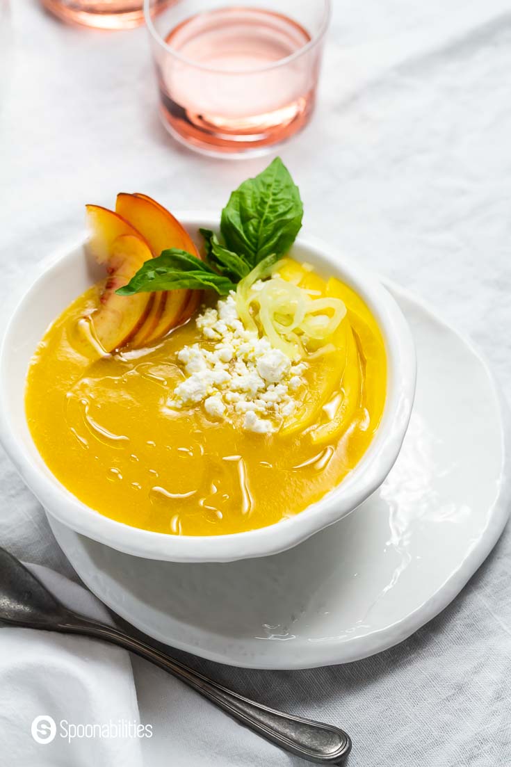 A side closes up of the chilled peach soup with goat cheese in a white bowl and tablecloth. Garnished with cucumbers, peaches, and yellow bell peppers. Recipe at Spoonabilities.com