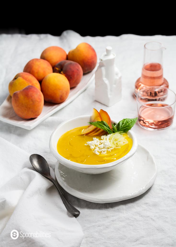 A table set up with a white linen tablecloth, in the center a white round plate, and on top a white bowl with a cold peach soup. Garnished with crumbled goat cheese, cucumber noodles, thinly sliced yellow bell pepper, and olive oil drizzle. Recipe at Spoonabilities.com