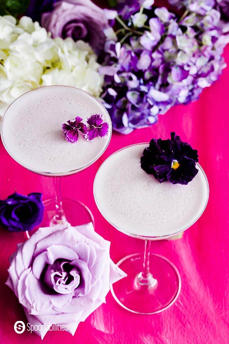 Overhead photo showing two glasses with the gin cocktail garnished with edible flowers. The drink has Creme de Violette for a beautiful purple/pink color. Recipe at Spoonabilities.com