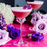 Close up photo with an elegant cocktail glass presenting the drink Gin Violette Cocktail garnished with edible flowers. In the background different flowers. Recipe at Spoonabilities.com